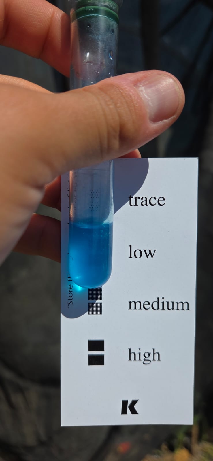 Hand holding a potassium (K) soil test tube with turquoise liquid beside a colour card used to read the nutrient level